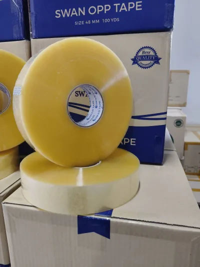 ม้วนเทปใสสองม้วนวางบนกล่องกระดาษ ด้านหลังเป็นกล่องเทปสวอนสีน้ำเงิน — SWAN TAPE โรงงานผลิตเทปกาว