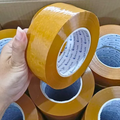 มือพนักงานถือม้วนเทปสีน้ำตาลเหนือกล่องที่บรรจุม้วนเทปเต็มกล่อง — SWAN TAPE โรงงานผลิตเทปกาว