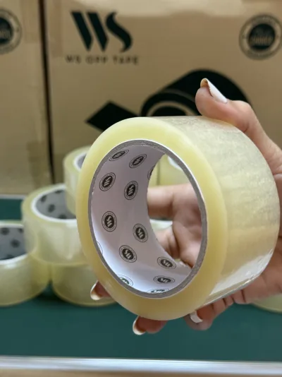 มือพนักงานถือม้วนเทปใสขึ้นให้ดู ด้านหลังเป็นกล่องเทปวางซ้อนกัน — SWAN TAPE โรงงานผลิตเทปกาว