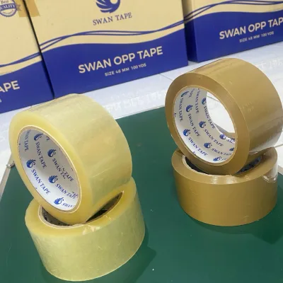 ม้วนเทปใสและน้ำตาลสี่ม้วนวางบนโต๊ะเขียว ด้านหลังเป็นกล่อง SWAN OPP TAPE — SWAN TAPE โรงงานผลิตเทปกาว