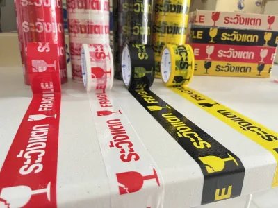 ม้วนเทประวังแตกสี่สีดึงเทปทาบบนโต๊ะขาว มีกล่องสินค้าอยู่ด้านหลัง — SWAN TAPE โรงงานผลิตเทปกาว