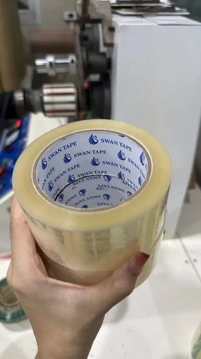มือถือม้วนเทปใส SWAN TAPE ที่เพิ่งตัดแบ่งเสร็จ ด้านหลังเป็นเครื่องตัดม้วนในโรงงาน — SWAN TAPE โรงงานผลิตเทปกาว