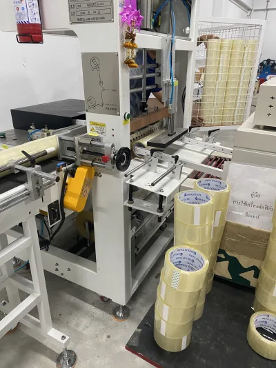 เครื่องตัดฟิล์มซีลเทปตั้งอยู่ในห้องผลิต มีม้วนเทปใส SWAN TAPE กองเรียงรออยู่ข้างเครื่อง — SWAN TAPE โรงงานผลิตเทปกาว